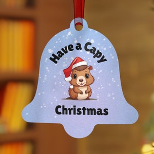 Op de afbeelding: Een klokvormige kerstversiering met een lichtblauwe achtergrond en een cartoon capibara met een kerstmuts. De versiering heeft de tekst "Have a Capy Christmas" en wordt opgehangen met een rood lint.