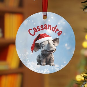 Op de afbeelding: Ronde kerstversiering met een rood lint. De versiering toont een cartoon neushoorn met een kerstmuts, met de naam "Cassandra" in het rood. De achtergrond is lichtblauw met sneeuwvlokken.