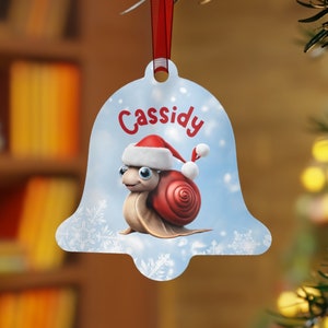 Op de afbeelding: Een klokvormige kerstversiering met een cartoon slak met een kerstmuts. De slak heeft een rood huisje en staat tegen een lichtblauwe achtergrond met sneeuwvlokken. De naam "Cassidy" staat in het rood.