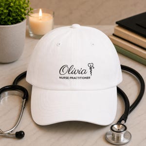 May include: A white baseball cap featuring the black printed text Olivia and NURSE PRACTITIONER, accompanied by a medical caduceus symbol with a heart shape at the top. The cap has a structured crown, a curved brim, and stitched eyelet details.