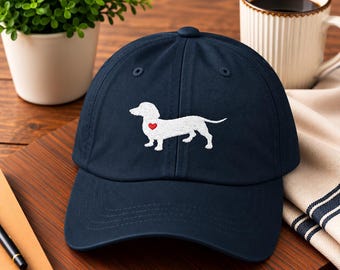 Puede incluir: Una gorra de béisbol azul marino con la silueta de un perro salchicha bordada en blanco en el panel frontal. El diseño del perro incluye un pequeño corazón rojo brillante en el pecho. La gorra tiene una corona estructurada y visera curva.