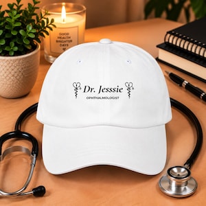 Könnte beinhalten: Eine weiße Baseballkappe mit dem schwarzen Schriftzug Dr. Jesssie OPHTHALMOLOGIST, flankiert von zwei schwarzen medizinischen Caduceus-Symbolen. Die Kappe hat eine strukturierte Krone, einen gebogenen Schirm und einen Knopf an der Oberseite.
