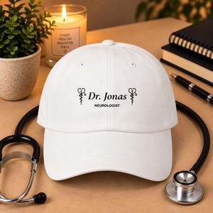 May include: A white baseball cap featuring black text that reads Dr. Jonas and NEUROLOGIST, centered between two black caduceus symbols. The cap has a structured crown, a curved brim, and a button at the top, presenting a clean and professional look.