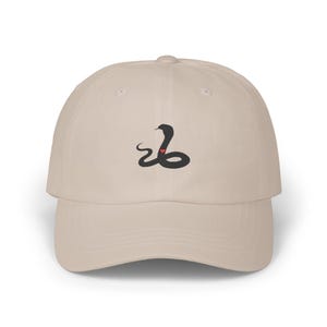 May include: A beige baseball cap with a black snake design and a red heart. The cap has a curved peak and a structured crown. The snake design is embroidered on the front.