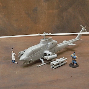 Pode incluir: Um modelo de helicóptero cinza com um design detalhado, apresentando um rotor e pods de mísseis. Figuras em miniatura de uma pessoa em jaleco de laboratório e um soldado são posicionadas perto, junto com uma pilha de mísseis. O modelo está em uma superfície marrom.