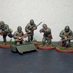 以下が含まれることがあります： オリーブドラブの制服、ヘルメット、ライフルを着た兵士のミニチュアフィギュア。フィギュアは、ひざまずいたり、狙いを定めたりするなど、さまざまなアクションポーズでポーズをとっています。小さな丸いベースに、シミュレートされた地形と小さな緑色の車両が取り付けられています。