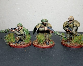 Könnte beinhalten: Drei Miniaturfiguren von Soldaten in Tarnuniformen, die jeweils ein Gewehr halten. Die Figuren stehen auf kleinen, runden Sockeln mit Grasbüscheln. Die Soldaten sind mit detaillierten Tarnmustern bemalt.