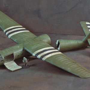 Puede incluir: Maqueta detallada de un avión de transporte militar verde oliva, con rayas negras y blancas en las alas y la cola. La puerta de carga está abierta y una figura de soldado en miniatura está cerca. Es un artículo de colección.