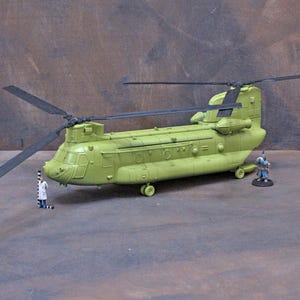 May include: A green Chinook helicopter model with black rotors. Two miniature figures are positioned near the helicopter. The model has a matte green finish, with detailed panel lines and rivets. The rotors are black and the wheels are visible.
