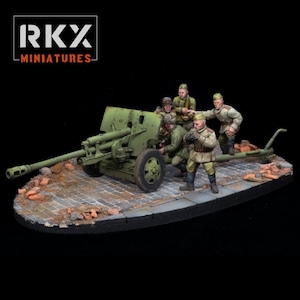 Può includere: Diorama in miniatura con un cannone di artiglieria verde con un gruppo di soldati in uniforme. La scena è ambientata su una base di ciottoli con detriti di mattoni. Il logo "RKX MINIATURES" si trova nell'angolo in alto a sinistra.