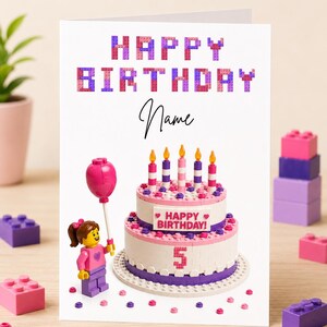 May include: A birthday card with a Lego-themed design. The card features the words "HAPPY BIRTHDAY" in block letters, a cake with candles, and a Lego figure holding a balloon. The number "5" is displayed on the cake.