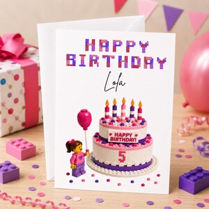 May include: A birthday card with the words "HAPPY BIRTHDAY Lola" in pixelated purple and pink letters. The card features a Lego-style cake with candles, a Lego figure, and a balloon. The number 5 is on the cake.