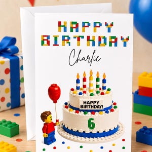 May include: A birthday card with a Lego theme, featuring the text "Happy Birthday Charlie". The card shows a cake with candles and a Lego figure with a red balloon. Colourful confetti and Lego bricks are scattered around the scene.