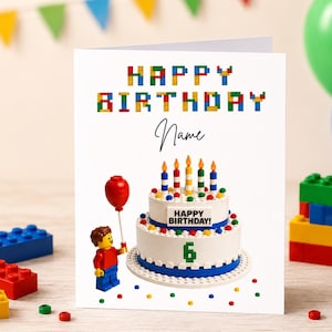 May include: A birthday card with a Lego-themed design. The card features the words "HAPPY BIRTHDAY" in colourful block letters. A cake with candles and the number 6 is on the card. A Lego figure holds a red balloon.