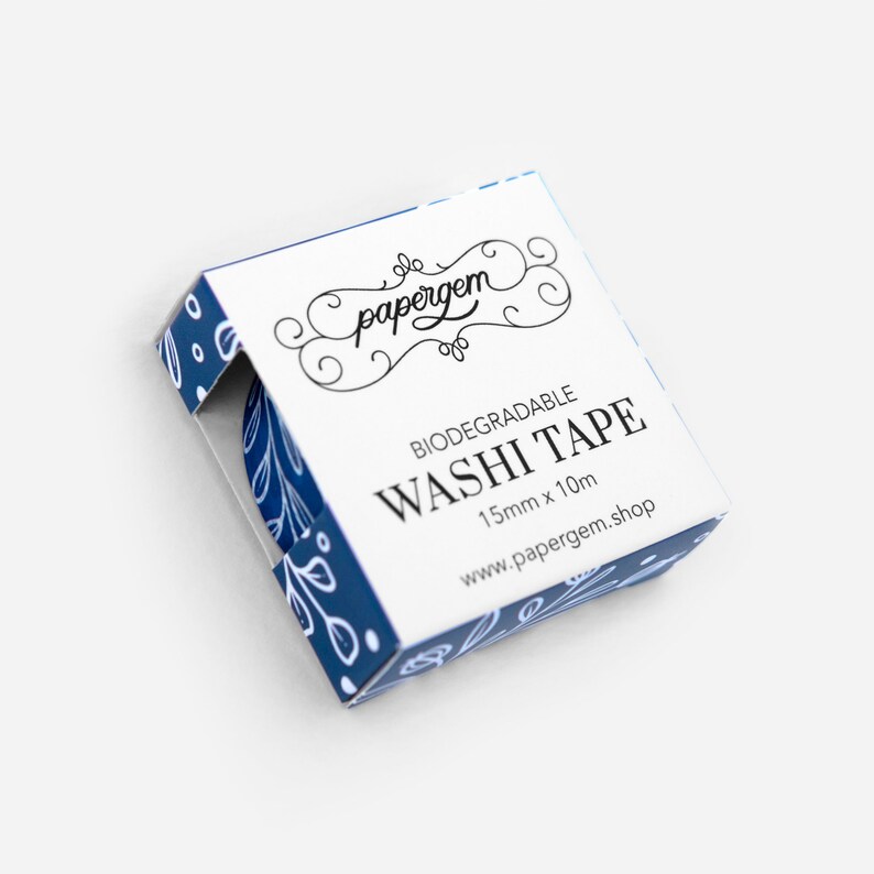 May include: A white box with blue floral pattern and the text "papergem" in a decorative font. The box contains a roll of biodegradable washi tape with a blue floral pattern. The text on the box reads "Biodegradable Washi Tape 15mm x 10m www.papergem.shop".