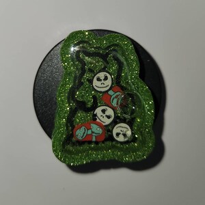 May include: Green glitter phone grip with a clear resin top featuring a Jack Skellington character from the Nightmare Before Christmas movie. The resin contains multiple Jack Skellington faces and a green and red creature.
