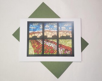 May include: Greeting card featuring a stained-glass window design. The artwork shows a landscape with a field of flowers, a mountain range, and a blue sky with clouds. The card is on a white background with a green envelope.