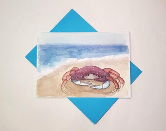 May include: Watercolour painting of a crab on a sandy beach with the ocean in the background. The crab is a deep red-purple colour with white claws. The painting is on a white card with a blue envelope.