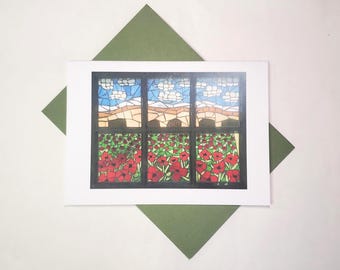May include: A white greeting card with a colourful illustration of a window scene. The window shows a landscape with blue skies, white clouds, and a field of red poppies. The card rests on a green envelope.