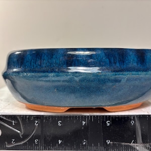 May include: A small, round bonsai pot with a glossy, deep blue glaze. The ceramic pot has a light brown base with three small feet. The pot's rim is slightly curved, and the overall shape is low and wide.