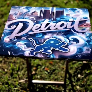 Könnte beinhalten: Ein Klapptisch mit dem Thema Detroit Lions. Der Tisch zeigt das Wort "Detroit" in einer stilisierten Schriftart, ein blaues Löwenlogo und ein Stadtbilddesign. Der Tisch steht im Freien auf einer grasbewachsenen Oberfläche.
