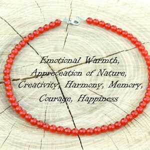 May include: A necklace made of small, round, red beads. The necklace is displayed on a light-colored, textured wooden surface. Text on the image reads: Emotional Warmth, Appreciation of Nature, Creativity, Harmony, Memory, Courage, Happiness.