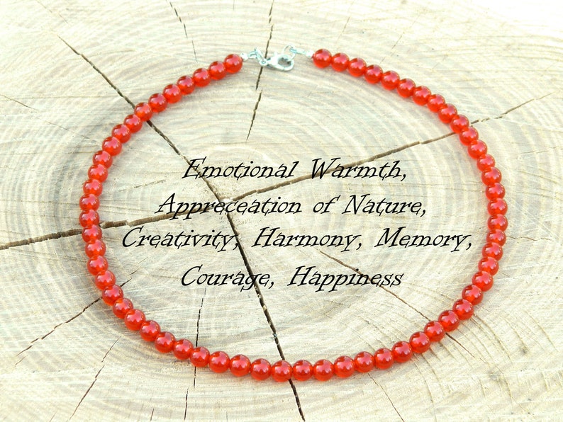 May include: A necklace made of small, round, red beads. The necklace is displayed on a light-colored, textured wooden surface. Text on the image reads: Emotional Warmth, Appreciation of Nature, Creativity, Harmony, Memory, Courage, Happiness.