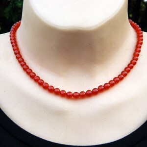 May include: A necklace of small, round, faceted red beads. The necklace is displayed on a mannequin, showcasing the jewelry's design and color. The beads are a vibrant shade of red.