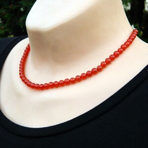 May include: A vibrant red beaded necklace. The necklace is composed of small, round beads, strung together to form a delicate strand. The necklace is displayed on a mannequin, against a black background.