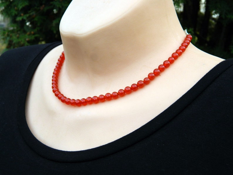 May include: A vibrant red beaded necklace. The necklace is composed of small, round beads, strung together to form a delicate strand. The necklace is displayed on a mannequin, against a black background.
