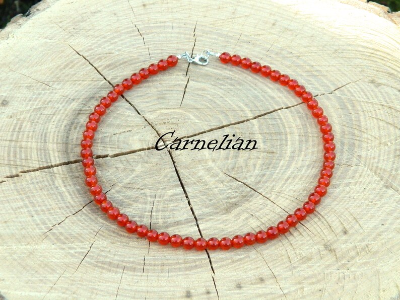 May include: A necklace made of small, round, orange-red carnelian beads. The necklace is displayed on a light brown, textured wooden surface. The word "Carnelian" is written in cursive.