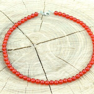 May include: A necklace composed of small, round, bright red beads. The beads are strung together, forming a complete circle. The necklace is secured with a silver-colored clasp. The background is a light-colored, textured wooden surface.