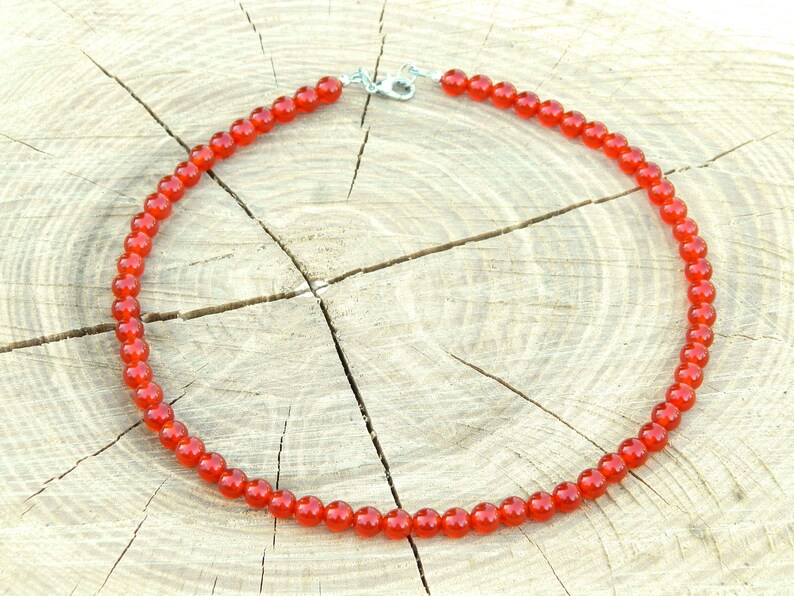 May include: A necklace composed of small, round, bright red beads. The beads are strung together, forming a complete circle. The necklace is secured with a silver-colored clasp. The background is a light-colored, textured wooden surface.