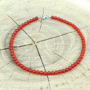 May include: A necklace featuring small, round, red beads strung together. The necklace has a silver-colored clasp and is displayed on a light brown, textured wooden surface. The beads are a vibrant, warm red color.