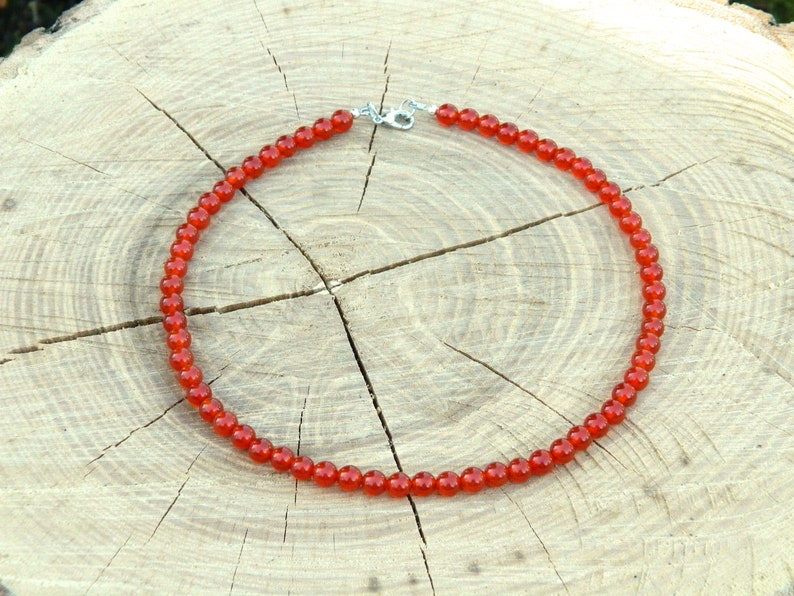 May include: A necklace featuring small, round, red beads strung together. The necklace has a silver-colored clasp and is displayed on a light brown, textured wooden surface. The beads are a vibrant, warm red color.