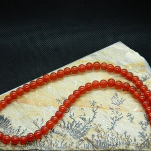 May include: A necklace composed of small, round, polished orange-red beads. The beads are strung together, forming a continuous strand. The necklace is displayed on a light-colored, textured stone surface against a dark background.