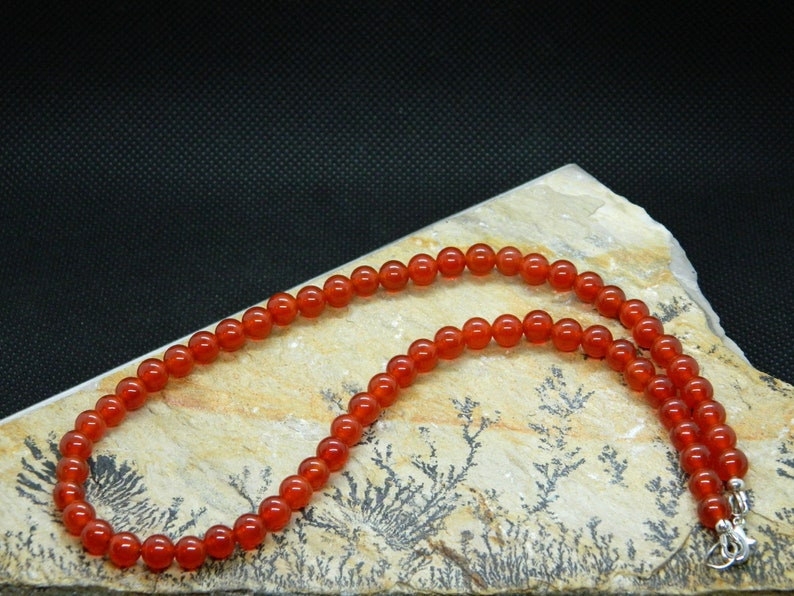 May include: A necklace composed of small, round, polished orange-red beads. The beads are strung together, forming a continuous strand. The necklace is displayed on a light-colored, textured stone surface against a dark background.