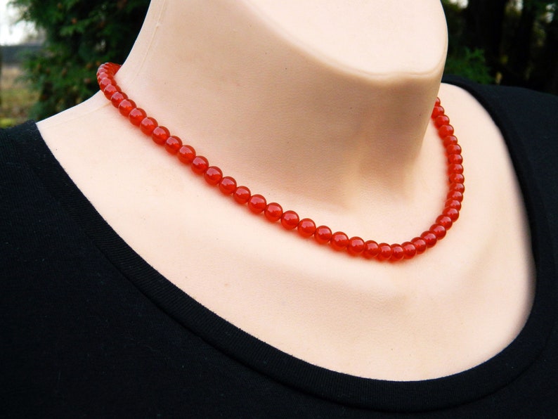 May include: A vibrant red beaded necklace. The necklace is composed of small, round beads, creating a continuous strand. The beads are a rich, saturated red color, and the necklace is displayed on a mannequin.