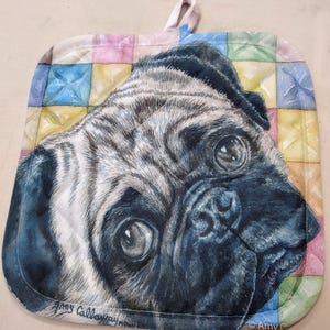 May include: A close-up of a pug's face on a colourful quilted pot holder. The pug has a light brown and black coat and is looking directly at the viewer. The pot holder has a patchwork design with squares of pink, blue, yellow, and green.