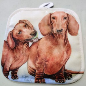 May include: Two brown dachshund dogs, one with a green ball in its mouth, are featured on a white pot holder with blue trim. The pot holder is decorated with a watercolor-style illustration of the dogs.