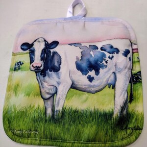 May include: A white and black cow with blue spots stands in a green field. The cow is looking at the viewer. The background is a light blue sky with a pink sunset. The image is on a white pot holder with a loop handle.