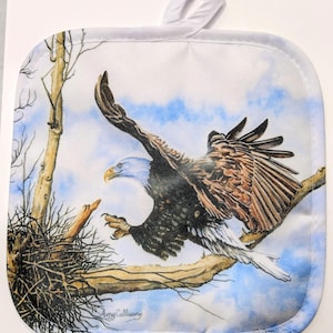 May include: A white cotton pot holder featuring a printed image of a bald eagle in flight. The eagle is perched on a branch with a nest below. The background is a blue sky with white clouds. The pot holder has a loop for hanging.