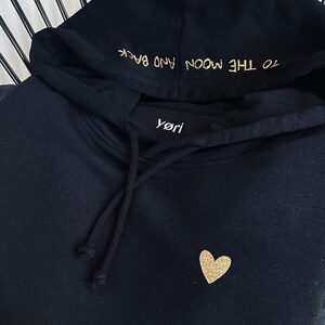 Könnte beinhalten: Schwarzer Hoodie mit einem goldenen Glitzerherz und dem Text "To the moon and back" auf der Kapuze.