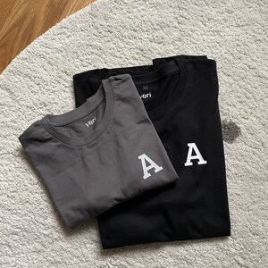 Könnte beinhalten: Zwei T-Shirts, eines grau und eines schwarz, mit einem weißen gestickten Buchstaben "A" auf jedem. Die Shirts sind gefaltet und übereinander gestapelt.
