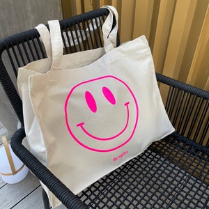 Könnte beinhalten: Eine beige Canvas-Tragetasche mit einem pinkfarbenen Smiley und dem Text "BE HAPPY" auf der Vorderseite.