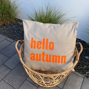 Könnte beinhalten: Ein beiger Kissen mit dem Text "hello autumn" in orangefarbenen Buchstaben liegt in einem geflochtenen Weidenkorb.