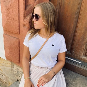 Könnte beinhalten: Ein weißes T-Shirt mit einem kleinen schwarzen Herz-Grafik, kombiniert mit einem hellrosa Rock. Die Person trägt eine runde Sonnenbrille und eine beige Umhängetasche. Eine goldene Halskette und Armbänder vervollständigen den Look. Im Hintergrund eine braune Tür und eine rotbraune Wand.