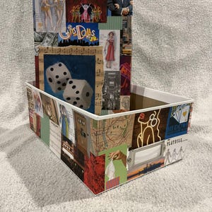 May include: A white cardboard box with a collage of images and text on the outside. The images include a map of New York City, a woman in a red dress, a pair of dice, and a musical score. The text includes the words "Playbill", "The Girl Dolls", and "Showboat".