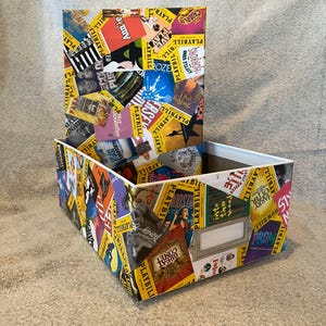 May include: A rectangular storage box and lid covered in colorful playbill covers. The box has a white interior and a metal label holder. The playbills feature various show titles and graphics.