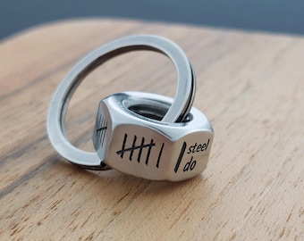 May include: A silver-colored keychain featuring a hexagonal nut-shaped charm. The charm has etched tally marks and the words "steel do." The keychain is on a wooden surface.